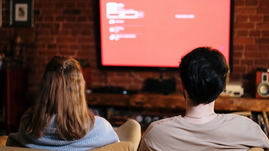 a couple sitting on a couch facing the TV. The screen is a red settings menu. 