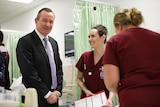 A smiling WA Premier Mark McGowan wearing a suit and tie stands in a hospital ward next to two student nurses in red scrubs.