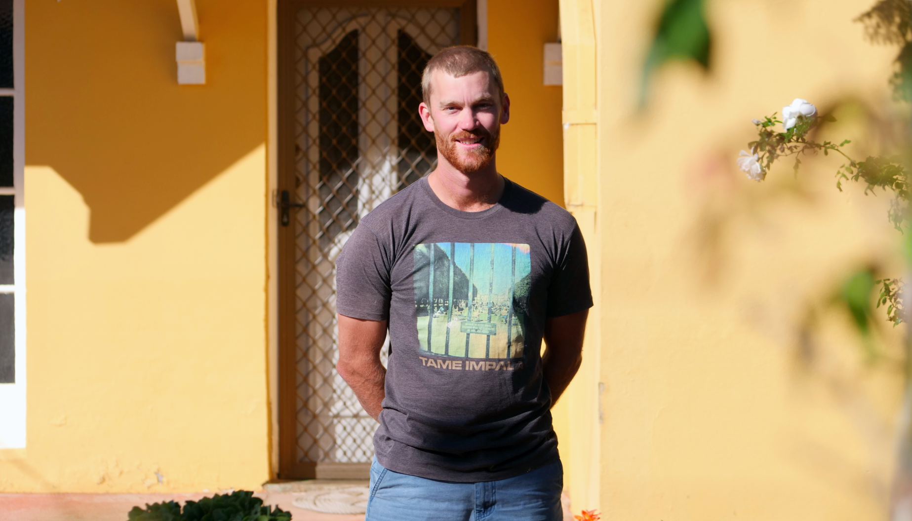 Man standing in front of yellow house smiles at the camera on sunny day.