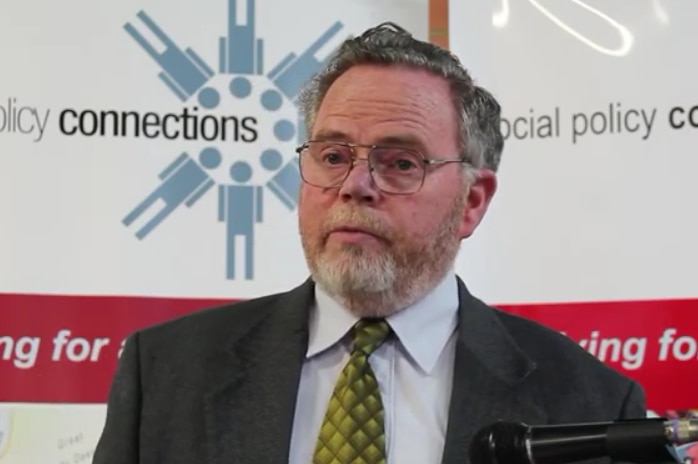 image of Caucasian man in a suit wearing spectacles with a microphone on the right hand side