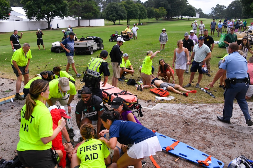 People lie on the ground at a golf course being attended to by others wearing fluorescent yellow shirts