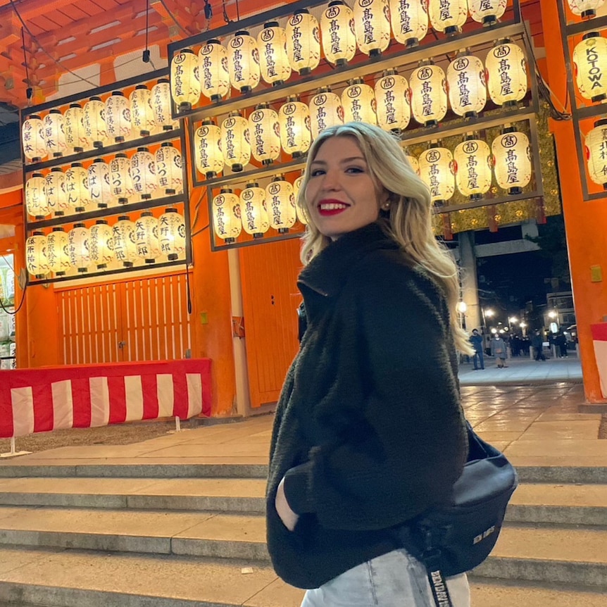 A blonde girl stands in front of some bright lanterns at night. 