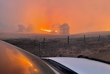 A bushfire burns in a paddock, as seen from behind a vehicle nearby.