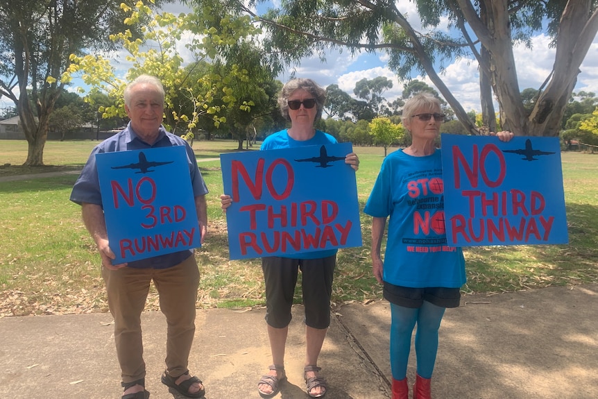 A man and two women hold signs saying "no third runway" in a sunny park.