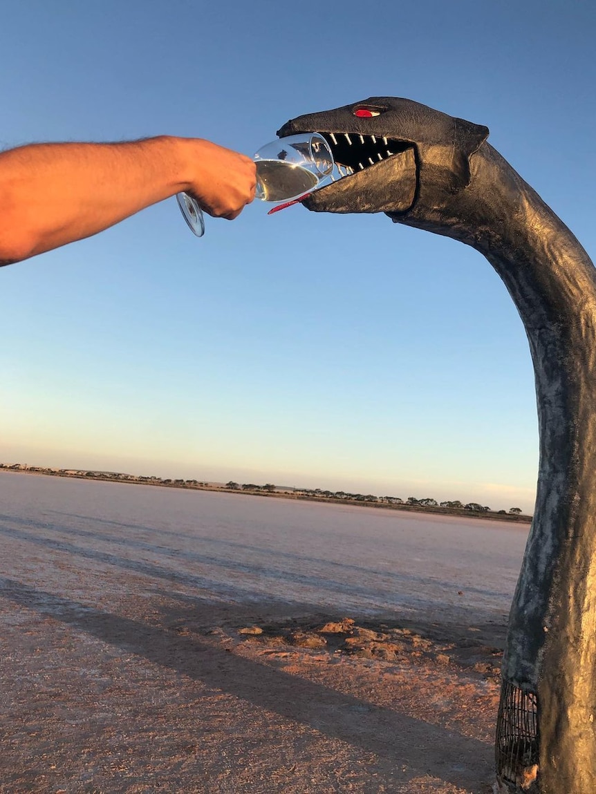 A person holds a glass of wine to the mouth of a metal sculpture shaped like the Lach Ness monster.