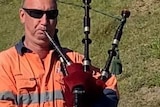 Man in fluro work gear plays bagpipes to a man and small child who wears a hat.