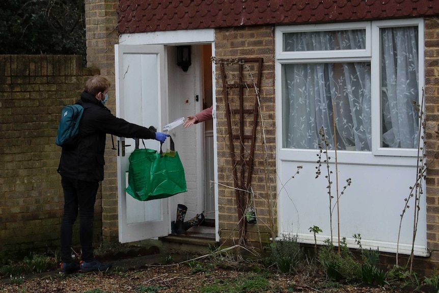 Man in coat and mask holds large green bag out toward open door