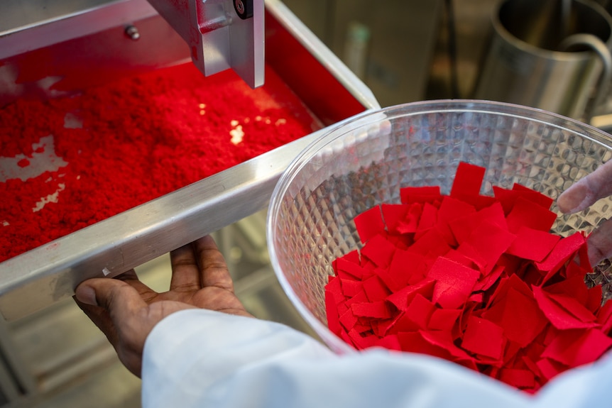 A scientist opens a draw from a machine showing crushed red clothing and hols a bowl of the original fabric in his other hand