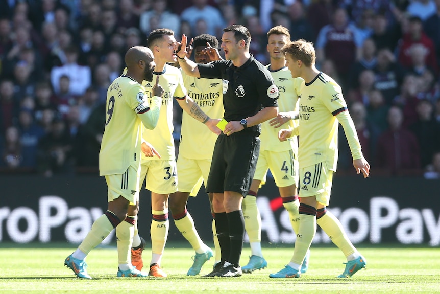 Arsenal players surround referee Andrew Madley, who has a yellow card in his hand 