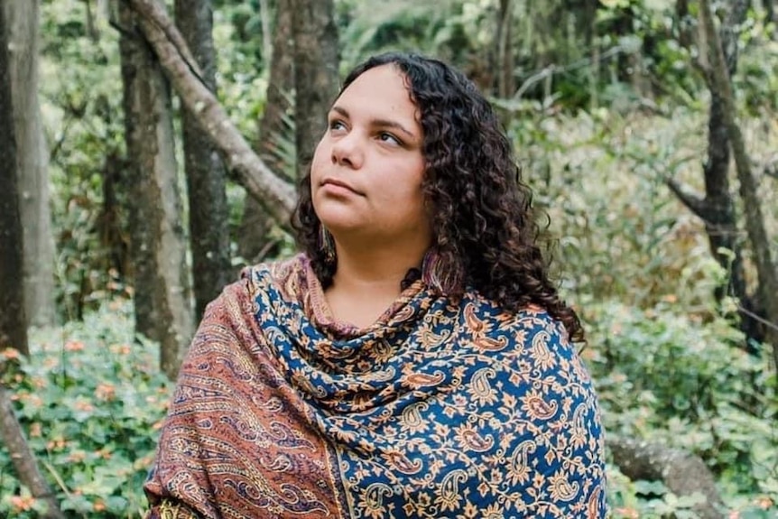 A woman looks to her left as she stands in a green forest. She wears a multicoloured shawl, her hair is down.