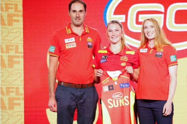 A young AFLW player holding up a jersey in between two other people in club colours.