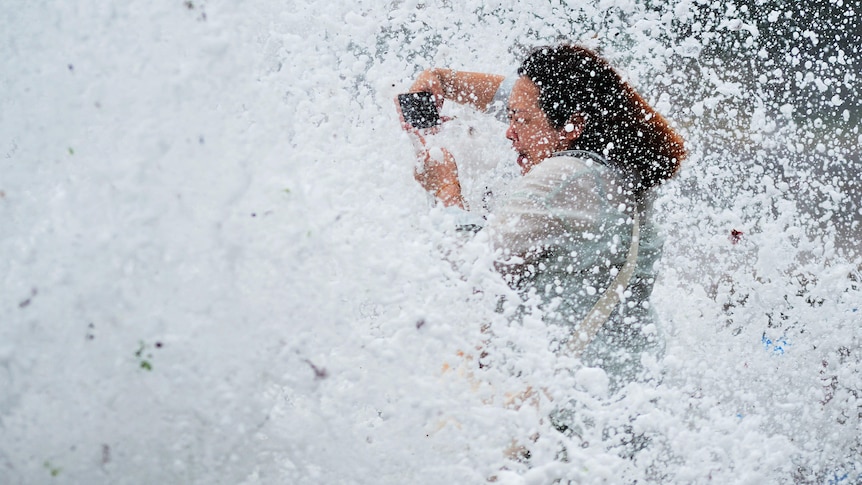 A woman holding a phone to take a photo hit by a wave. 