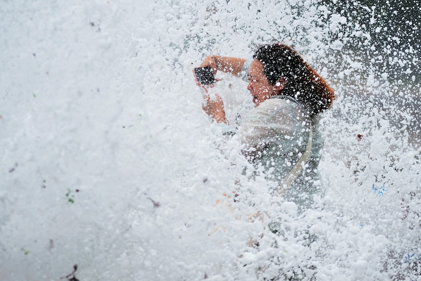 A woman holding a phone to take a photo hit by a wave. 