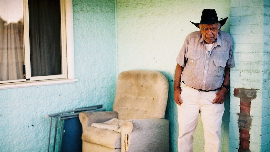 An elderly Aboriginal man in a black cowboy hat leans against a green wall.