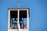 A priest holds up the eucharist while standing in a belltower against a blue sky.