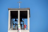 A priest holds up the eucharist while standing in a belltower against a blue sky.