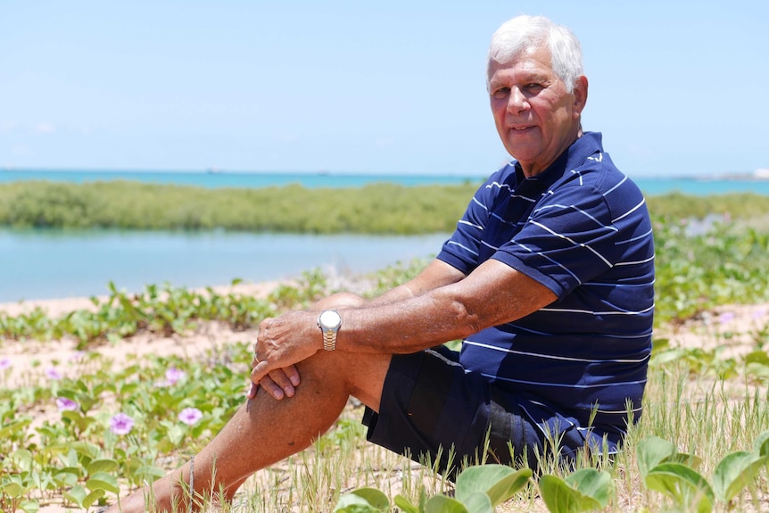 Man with grey hair sits on a beach in the sun.