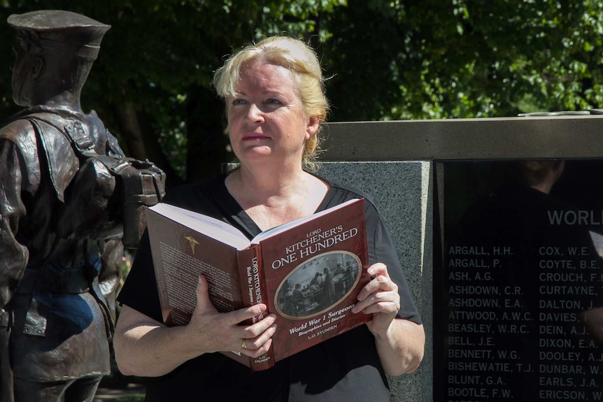 A woman holds a book called Lord Kitchener's One hundred in front of a war memorial