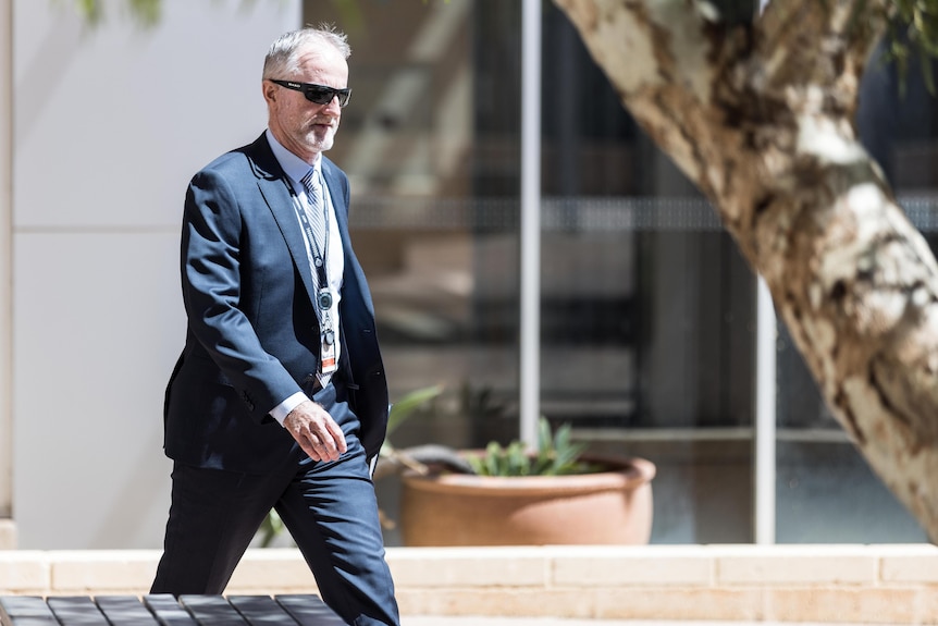 A bearded, grey-haired man in a dark suit and sunglasses walks away from a court building on a sunny day.