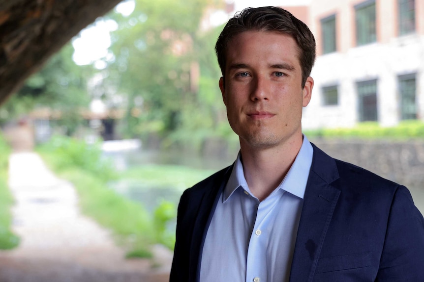 A tall, young and handsome white man stands under a bridge in a blue suit with a leafy river behind.