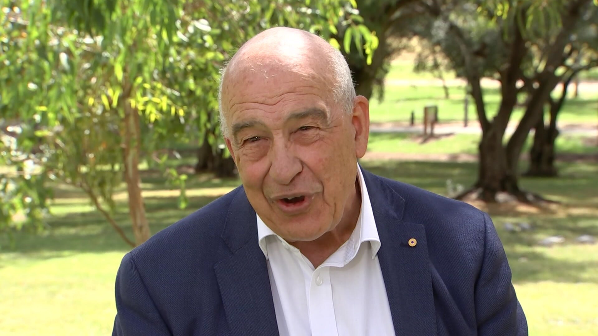 A man in a suit speaks at a park with trees