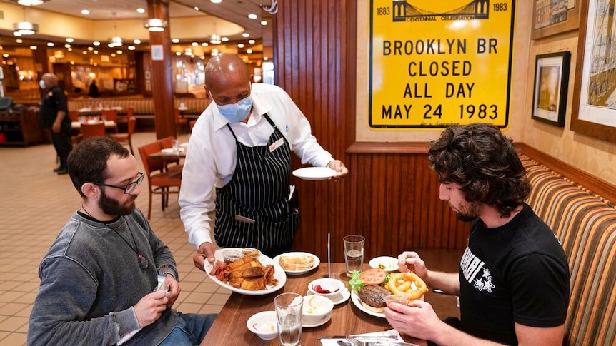 Waiter wearing a mask serves lunch to two men at Junior's Restaurant in New York.