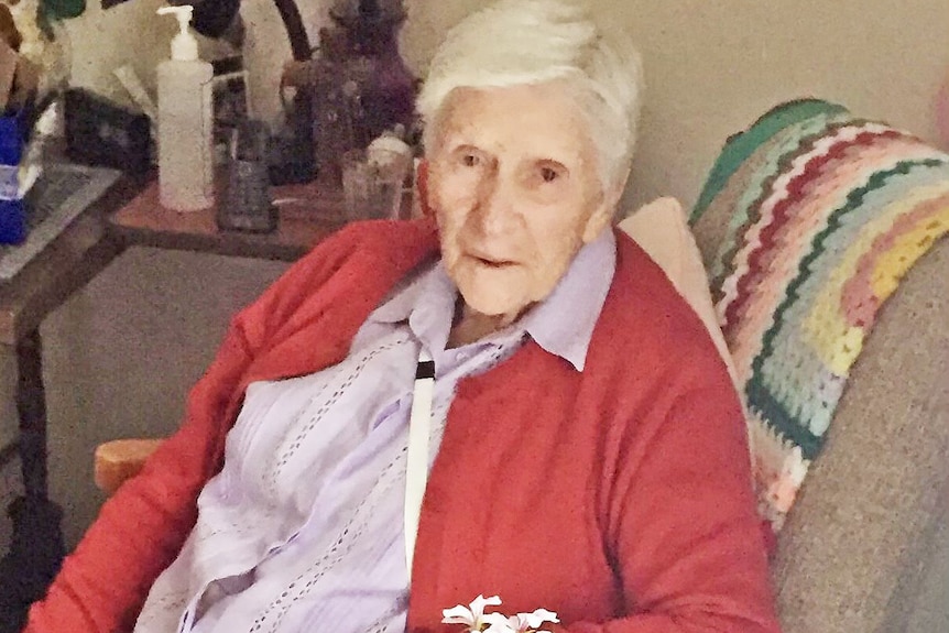 elderly woman wearing a red cardigan and sitting in an armchair in front of a pot with flowers, looking up at the camera