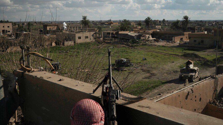 A fighter stands atop a building holding a mounted machine gun overlooking a village.