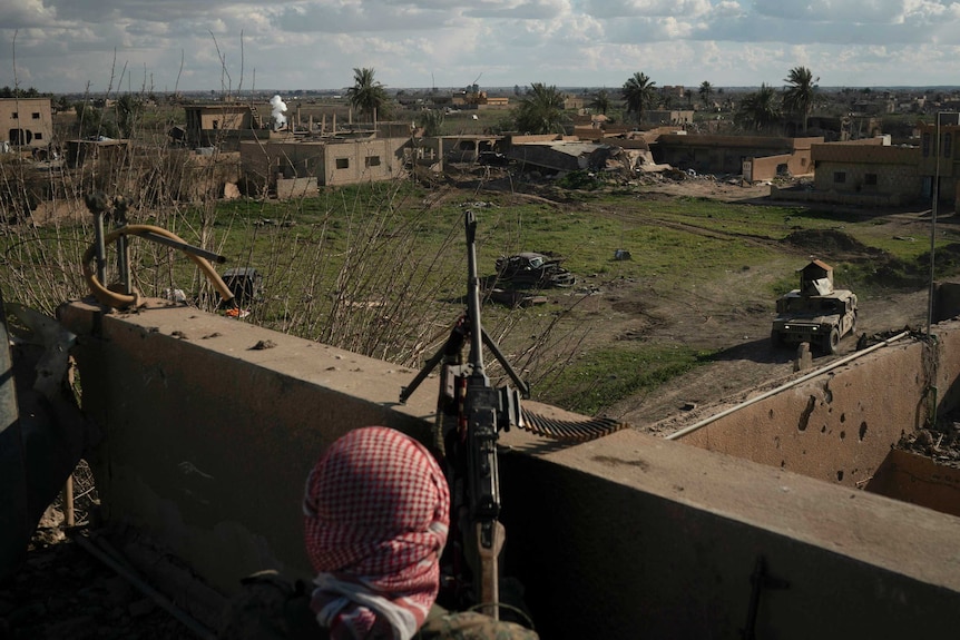 A fighter stands atop a building holding a mounted machine gun overlooking a village.
