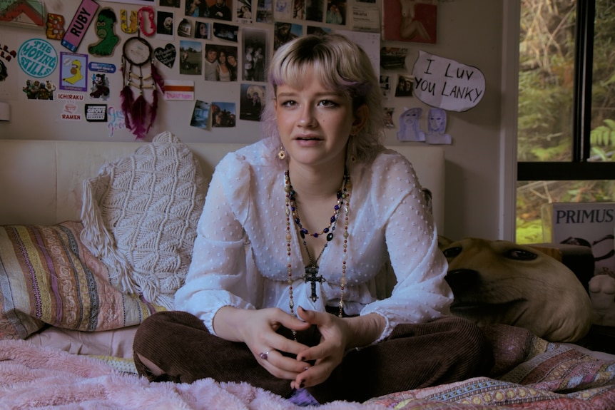 A teenaged girl sits on her bed with a wall of photos behind her, wearing beads and ends of hair dyed pink