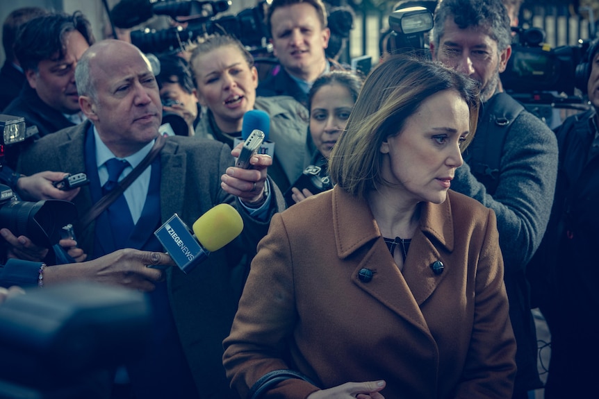 A film still of Keeley Hawes being swamped by reporters brandishing microphones in her face.
