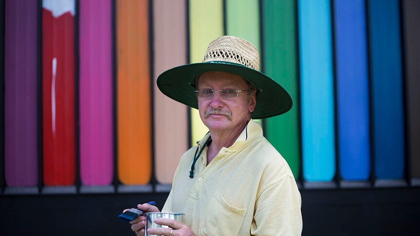Bill Gannon stands holding a pot of paint and brush, with rainbow colours in background
