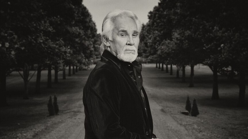 Black and white photo of Kenny Rogers standing on a tree-lined driveway