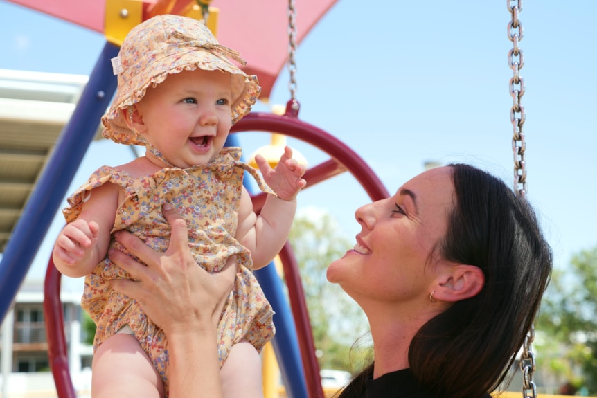 Janita looks up smiling at her daughter who she's holding up in her hands at a playground.