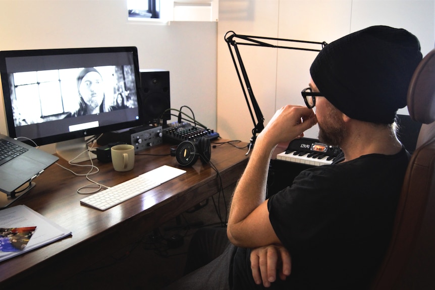 A man wearing a beanie and glasses sits at his laptop.