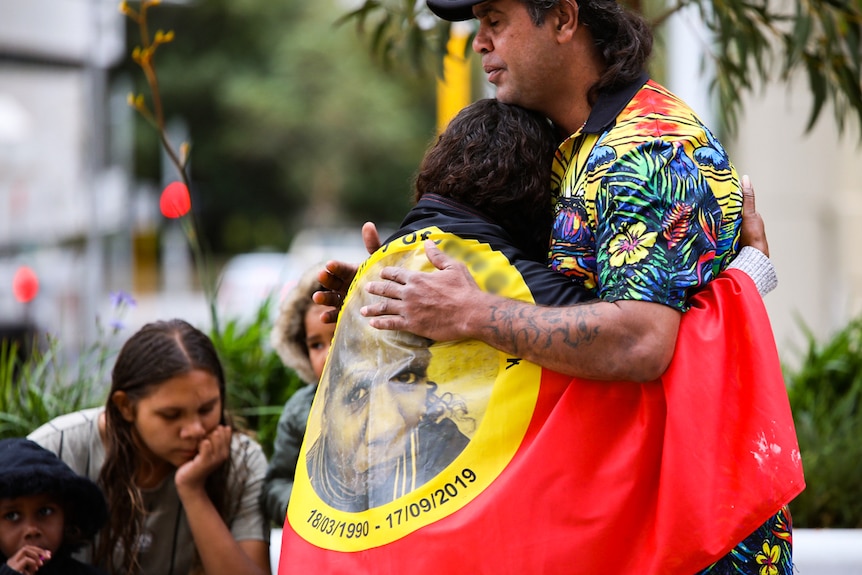 A man hugs a woman who is wearing an Aboriginal flag over her shoulders.
