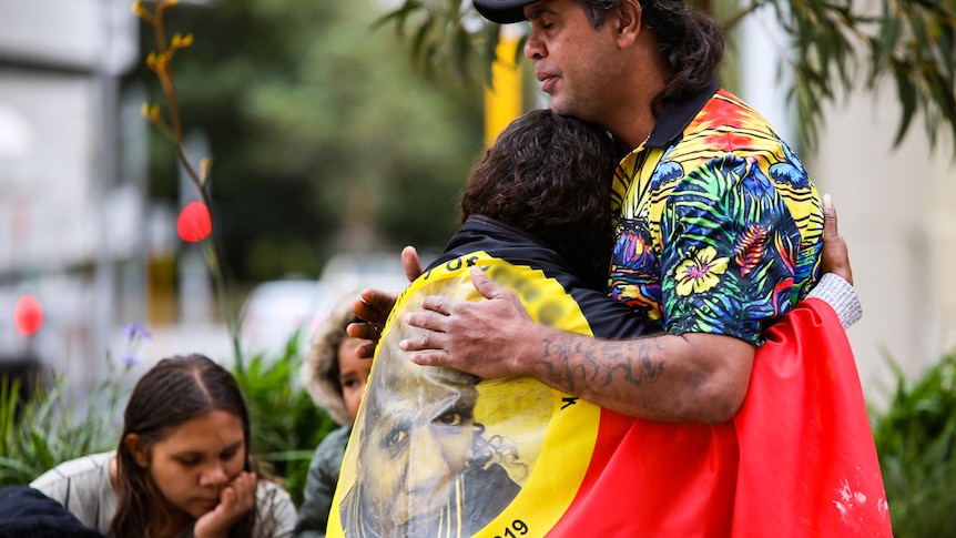 A man hugs a woman who is wearing an Aboriginal flag over her shoulders.