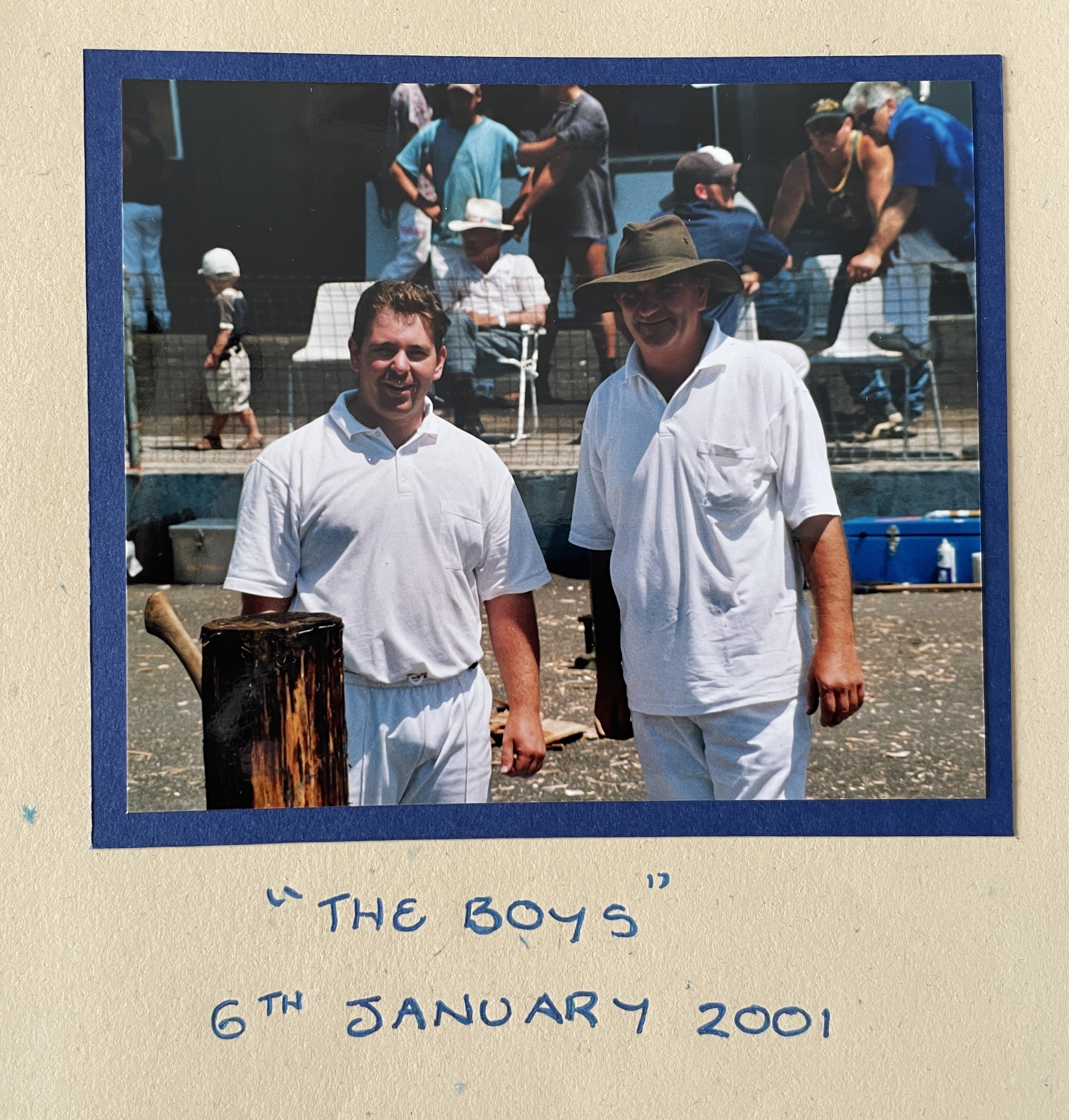father and son in white tops and pants stand together, with the son holding an axe next to a log.