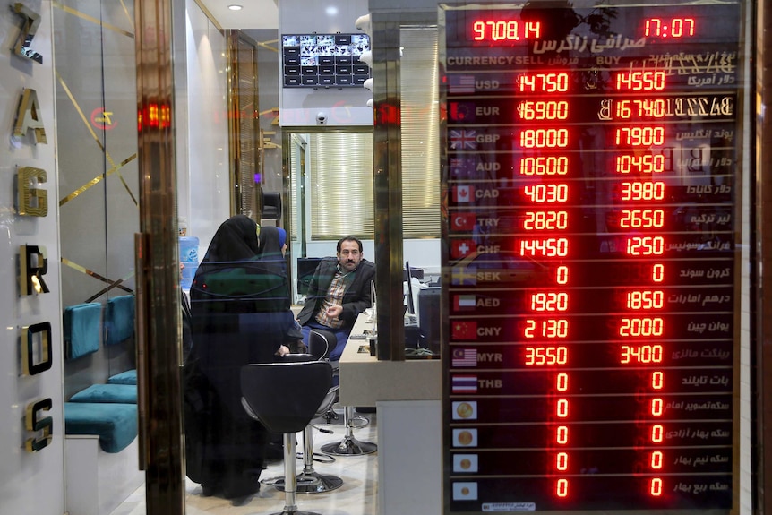 Customers speak with a money exchanger at his shop in Tehran. The board showing Iran's poor exchange rates is visible.