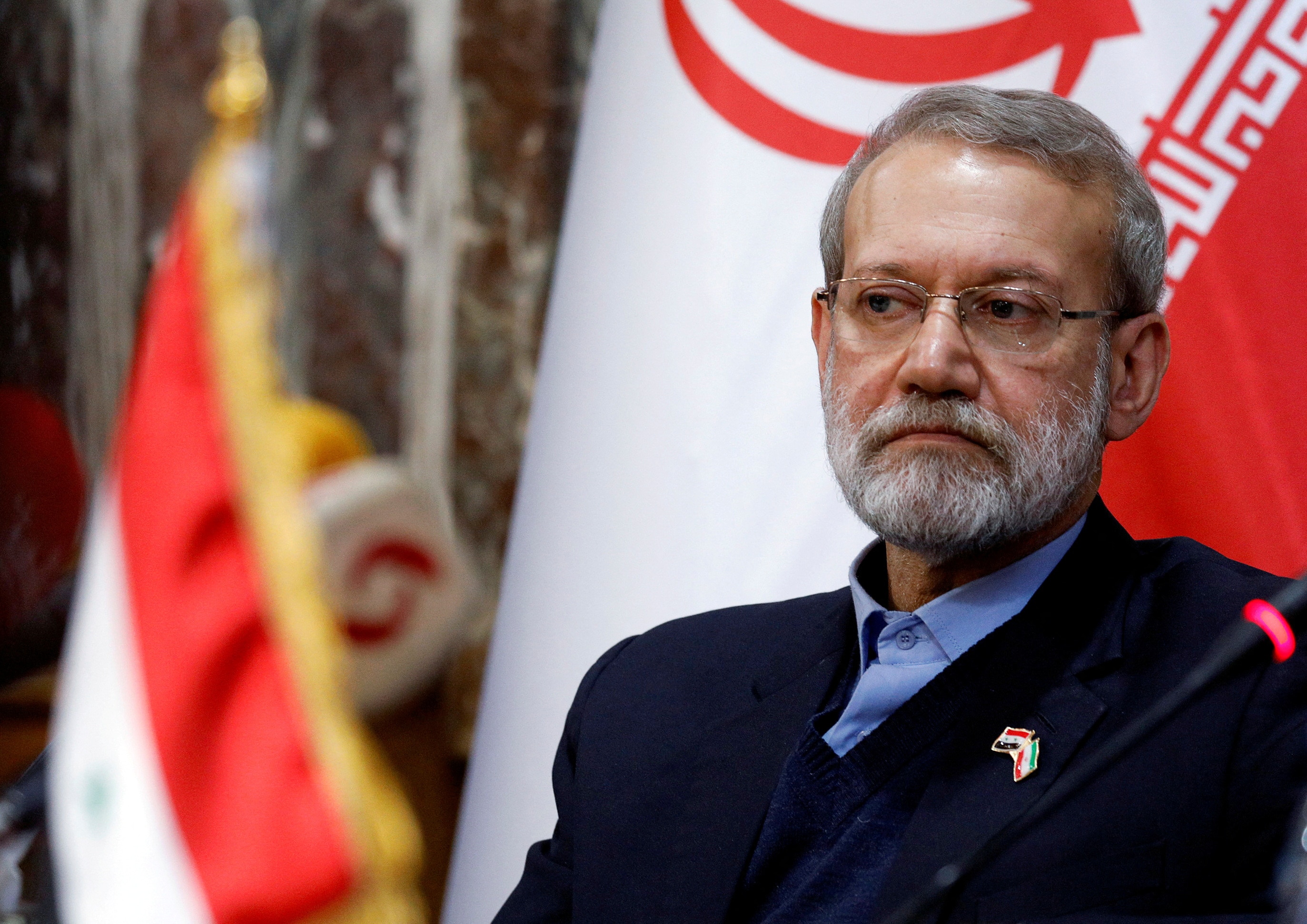 Ali Larijani wearing a dark suit sits in front of an Iranian flag with a serious expression.