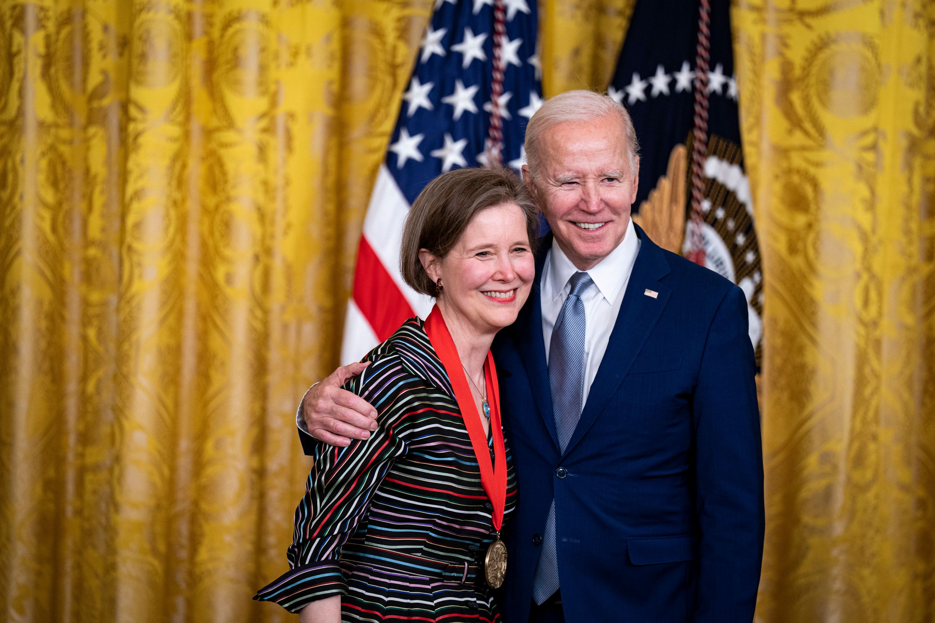 President Joe Biden with his arm around Ann Patchett, who has a medal around her neck, the US flag behind them.