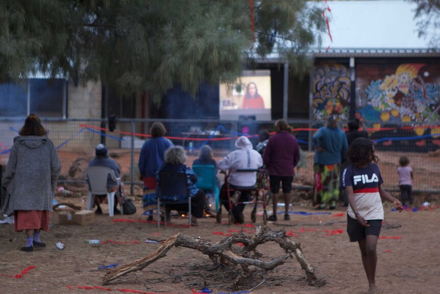 People sitting on chairs in the open, watching a screening of the national indigenous art awards