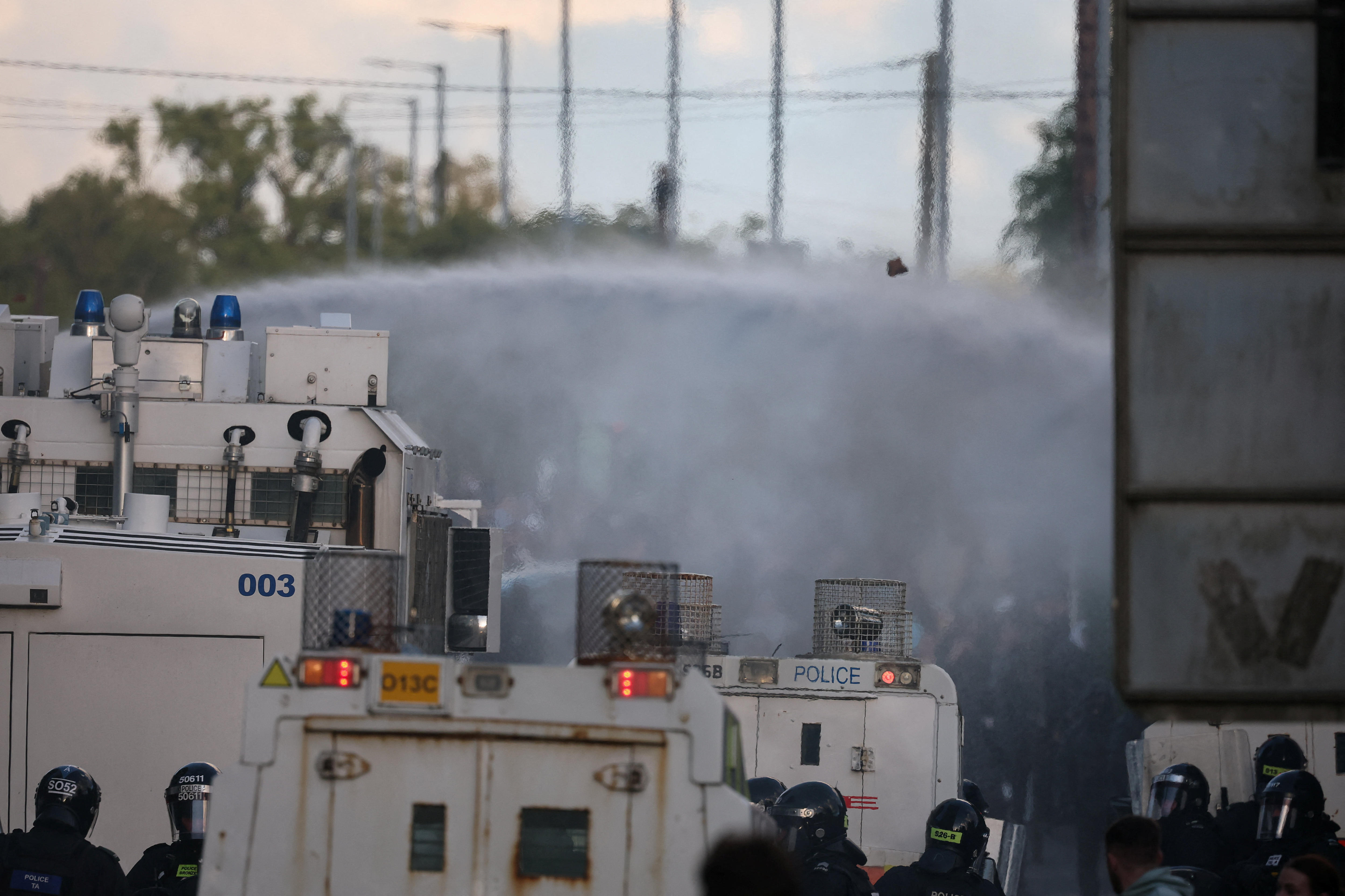 Armoured police stand near an armoured police vehicle spraying water.