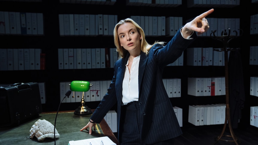 A young, smartly dressed blonde woman has one hand on a desk, the other pointing in the air