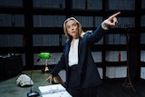 A young, smartly dressed blonde woman has one hand on a desk, the other pointing in the air
