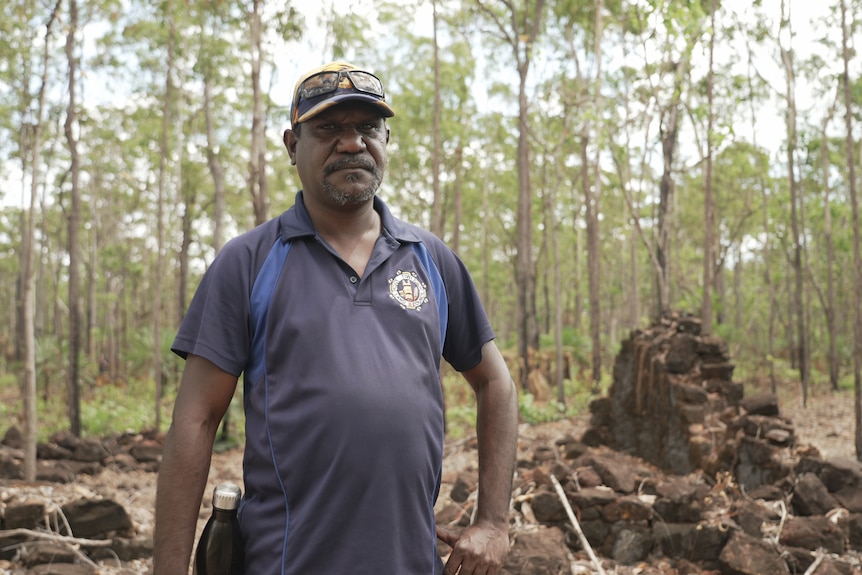 An Indigenous man standing in a forest with remnants of a brick building behind him
