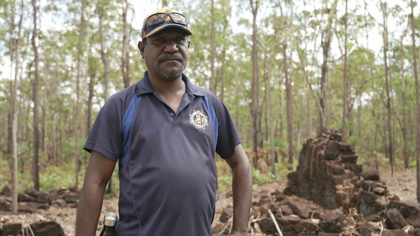 An Indigenous man standing in a forest with remnants of a brick building behind him