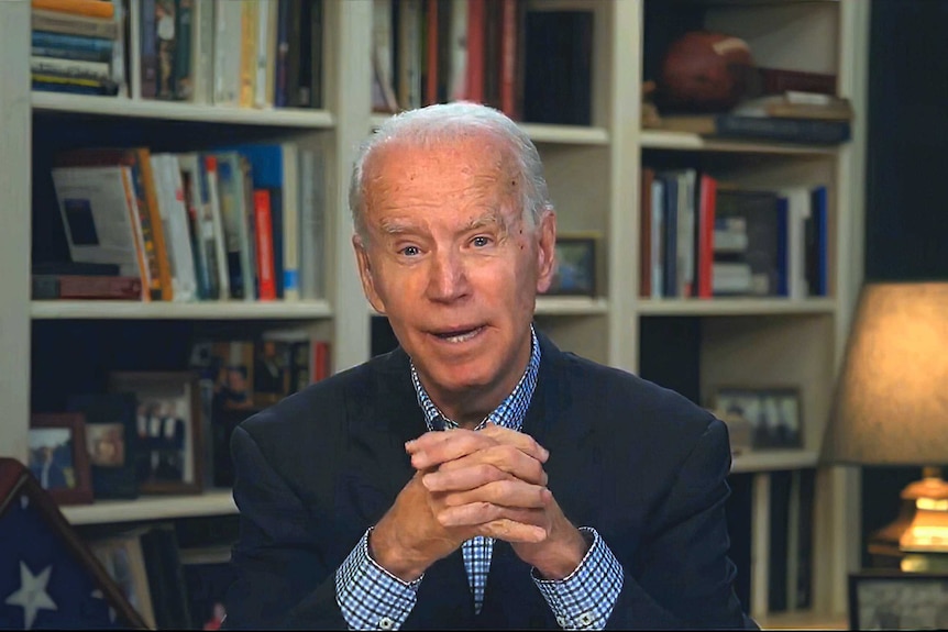 Joe Biden, sitting in front of a bookshelf, looks at the camera as he speaks.