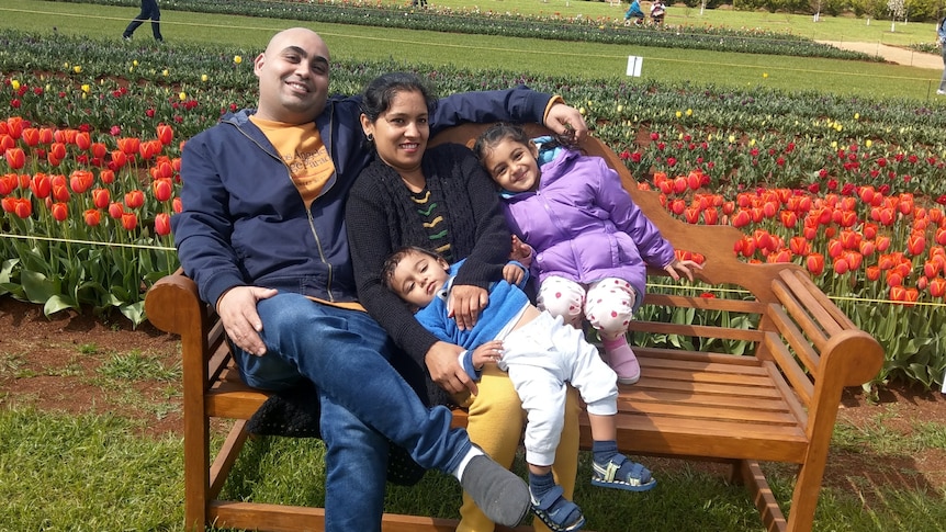 A woman, man, girl and boy smile and sit closely together on a bench as they post for a photo in a flower garden.
