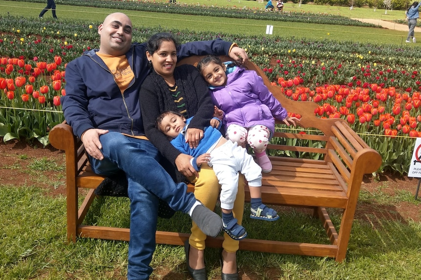 A woman, man, girl and boy smile and sit closely together on a bench as they post for a photo in a flower garden.